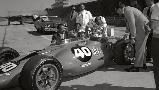Andy Granatelli, behind cockpit in suit, and his brothers, Vince, left in STP uniform, and Joe, right in STP uniform, help Johnny Carson get comfortable in the STP Turbine in 1967 at the Indianapolis Motor Speedway. Driver Parnelli Jones is behind them in a white firesuit.